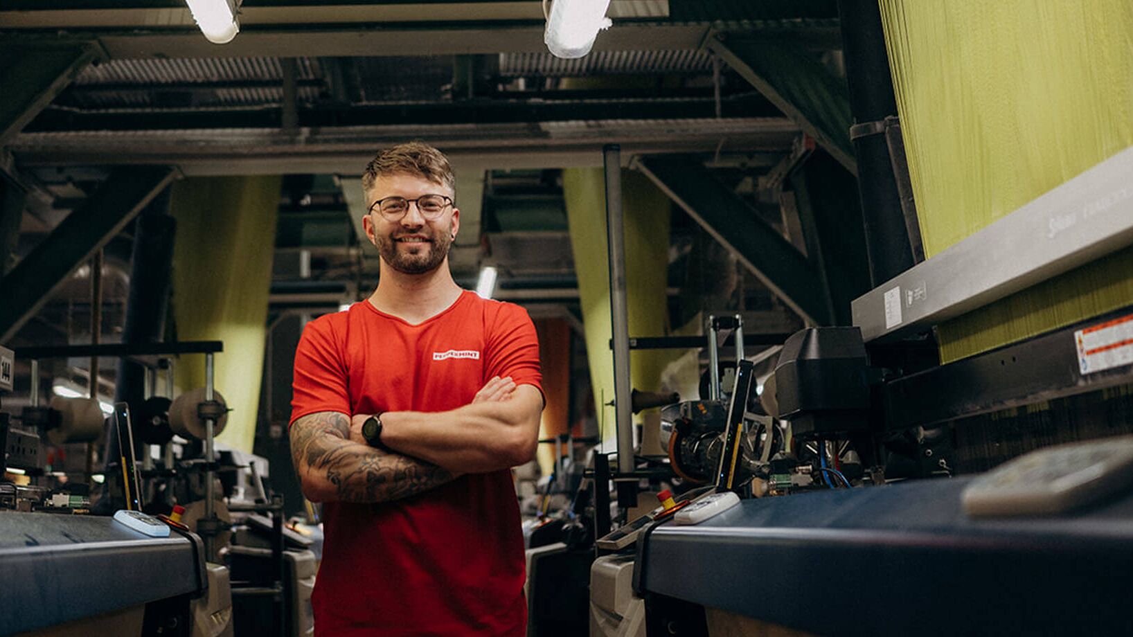 A man in a red shirt stands confidently with crossed arms in an industrial setting, showcasing advanced machinery.