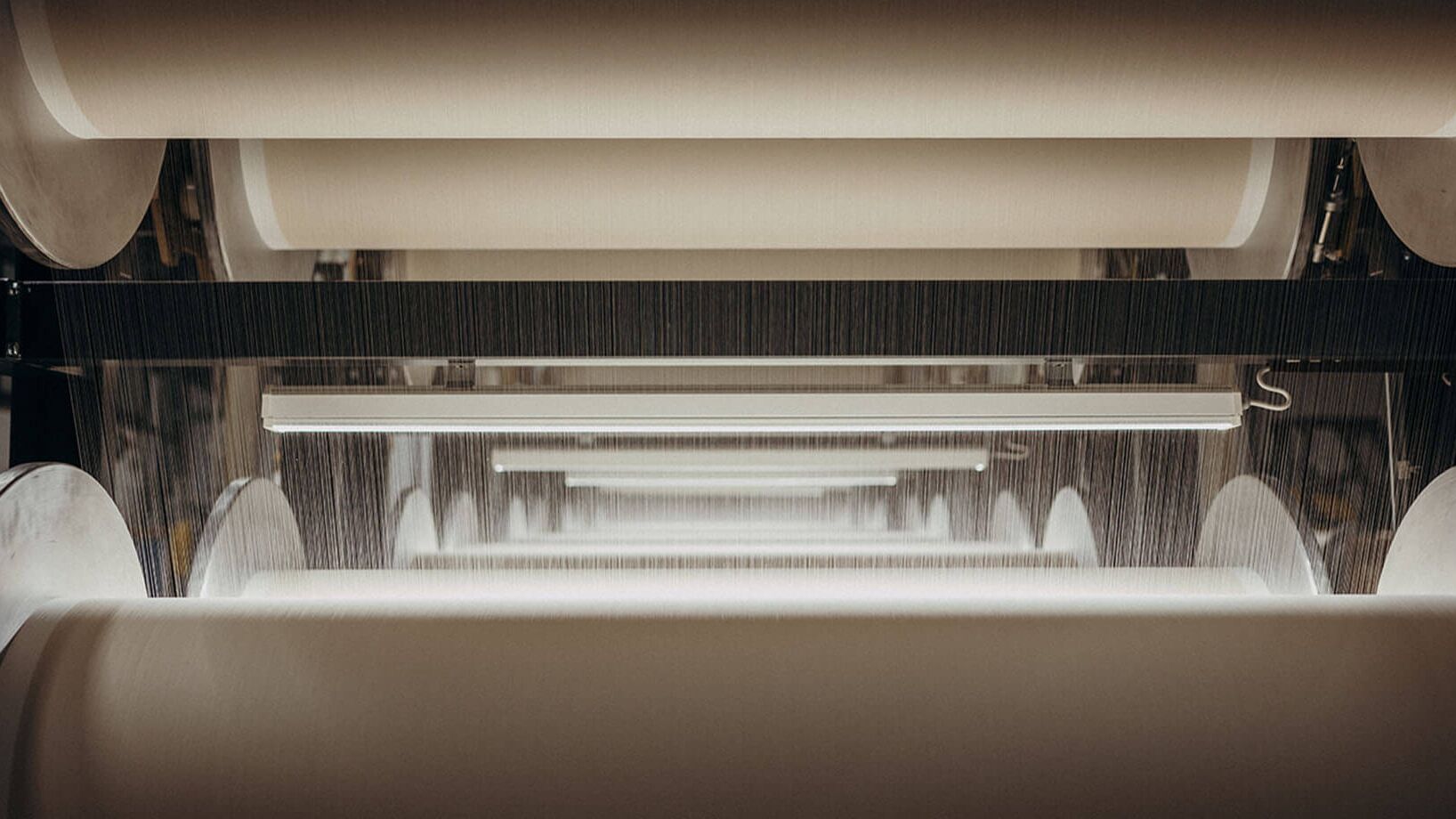 Large industrial textile rollers are aligned in a factory, with fabric being processed under bright lights.