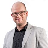 A man in a grey blazer stands confidently with hands in pockets against a plain white background.