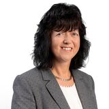 A woman in a gray blazer smiles confidently against a plain white background.